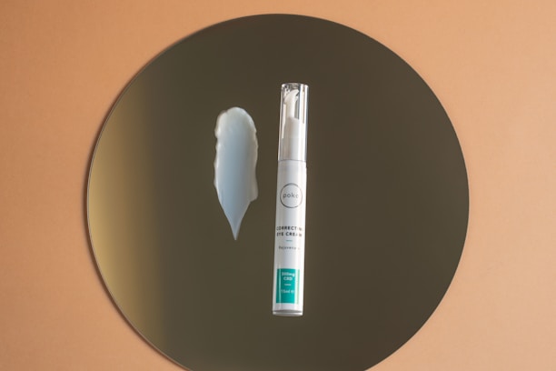 A small bottle of eye cream with a pump dispenser is placed upright on a circular reflective surface, possibly a mirror, against a warm brown background. Next to the bottle, there is a smear of white cream on the mirror, illustrating the texture of the product.