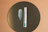 A small bottle of eye cream with a pump dispenser is placed upright on a circular reflective surface, possibly a mirror, against a warm brown background. Next to the bottle, there is a smear of white cream on the mirror, illustrating the texture of the product.