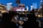 Two people sit in a convertible car at a rooftop drive-in theater. The city skyline is visible in the background, with several skyscrapers illuminated against a deep blue evening sky. The movie screen ahead displays credits featuring names such as Tom Guiry and Mike Vitar.