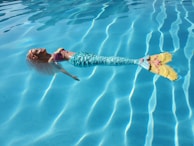 A mermaid doll floats on the surface of a swimming pool. The doll is dressed in a colorful bikini top and a shimmery, patterned turquoise mermaid tail with yellow and pink accents at the fin. The water is clear, creating a wavy reflection pattern.