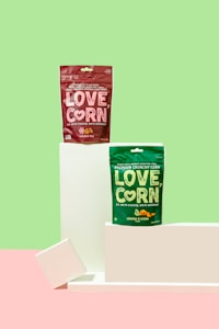 Two packages of LOVE, CORN snacks are displayed against a bright, pastel background. The red package is labeled as 'smoked bbq' flavor, while the green package is 'cheese & onion' flavor. They are positioned on minimalist white podiums, enhancing the clean and modern look.