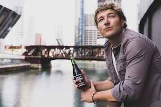 A person is leaning on a railing by a river in an urban setting, holding a bottle labeled 'Sangria Senorial'. The background features a cityscape with modern buildings and a bridge over the water.