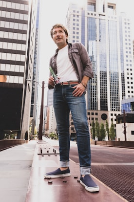 A person stands confidently on a city sidewalk, surrounded by tall, modern skyscrapers. They are casually dressed in jeans, a white t-shirt, and a button-up shirt. The setting is urban, with large glass buildings towering overhead.
