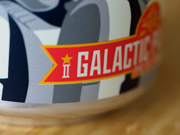 A close-up view of a product label featuring bold, white text that says 'GALACTIC' on a red banner with a yellow border. A small star and the Roman numeral 'II' are also present. The background includes gray and black abstract shapes. The texture of the label appears smooth and glossy.