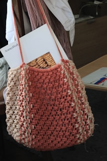 A crocheted bag in shades of salmon pink and beige hangs from a chair. Inside the bag, there is a magazine with an image of baked goods on its cover. The background includes a white garment draped over the chair and some indistinct objects on a desk.