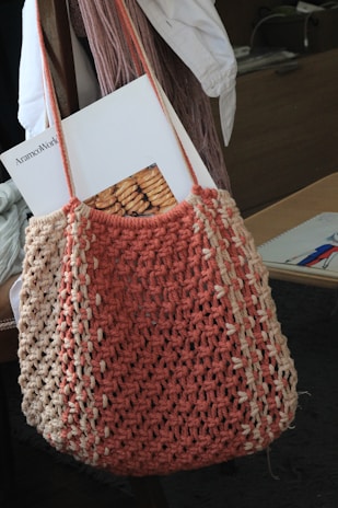 A crocheted bag in shades of salmon pink and beige hangs from a chair. Inside the bag, there is a magazine with an image of baked goods on its cover. The background includes a white garment draped over the chair and some indistinct objects on a desk.