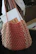 A crocheted bag in shades of salmon pink and beige hangs from a chair. Inside the bag, there is a magazine with an image of baked goods on its cover. The background includes a white garment draped over the chair and some indistinct objects on a desk.