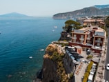 A cliffside hotel with a red facade overlooks a wide expanse of blue sea, with a backdrop of mountains in the distance. The architecture features balconies and a rooftop terrace with lounge chairs. Small boats are scattered on the calm water near the cliffs, and lush greenery surrounds the hotel.