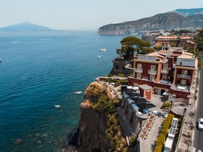 A cliffside hotel with a red facade overlooks a wide expanse of blue sea, with a backdrop of mountains in the distance. The architecture features balconies and a rooftop terrace with lounge chairs. Small boats are scattered on the calm water near the cliffs, and lush greenery surrounds the hotel.