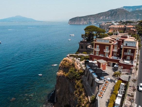A cliffside hotel with a red facade overlooks a wide expanse of blue sea, with a backdrop of mountains in the distance. The architecture features balconies and a rooftop terrace with lounge chairs. Small boats are scattered on the calm water near the cliffs, and lush greenery surrounds the hotel.