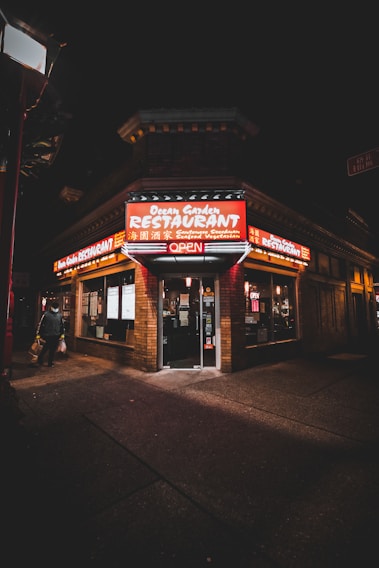 A corner restaurant with a brick facade is illuminated by bright red and yellow neon signs. The signage indicates that it is open and offers Cantonese, Szechuan, seafood, and vegetarian dishes. The dark night sky contrasts with the warm light from the restaurant. A person is walking away from the restaurant, carrying a bag.