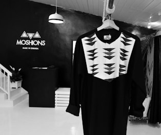 A black long-sleeved garment with a geometric white and black pattern on the chest area hangs from a wooden hanger. The background features a minimalist store interior with a black wall and white flooring. The brand logo 'MOSHIONS' with the tagline 'Made in Rwanda' is displayed on the wall. A staircase and a potted plant are visible to the left.