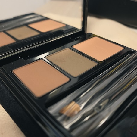 An open makeup palette features three shades of powder, accompanied by a brush, all enclosed in a glossy black case. The colors include two shades of brown and a neutral olive, with a reflective mirror on the inside of the lid. A soft, warm lighting casts gentle shadows over the palette, enhancing the earthy tones.