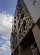 A modern building with a unique, perforated facade featuring various circular patterns. The facade creates interesting shadows on the walls, and the building has several windows. A sign with the word 'HOTEL' is prominently displayed. The sky is partly cloudy, with patches of blue visible.