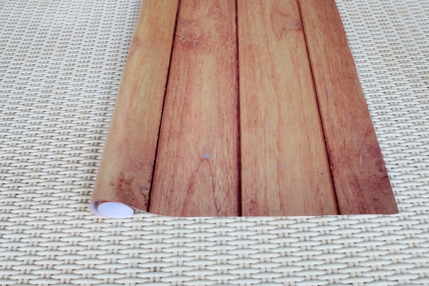 A piece of paper or material with a wooden pattern print is rolled partially on one side, placed on a surface with a woven pattern.