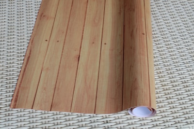 A rolled sheet of light wood-patterned wallpaper is laid out on a woven, off-white surface. The wallpaper features vertical lines and resembles wooden planks.