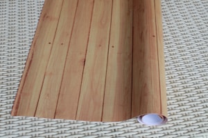 A rolled sheet of light wood-patterned wallpaper is laid out on a woven, off-white surface. The wallpaper features vertical lines and resembles wooden planks.