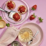 Three small strawberry tarts are placed on a glass plate with a metal serving utensil, surrounded by loose strawberries and tart shells on a pink textured surface. A cup of tea sits on a pink plate with a spoon beside it, accompanied by a light pink napkin and a tea infuser containing loose tea leaves.