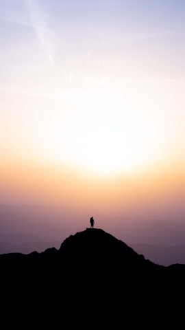 A sunset silhouette of a man standing tall on a mountain peak after a climb.