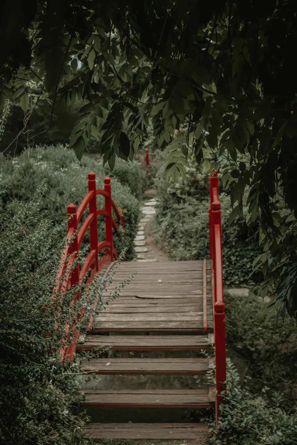 Red wooden bridge with curved pillars spans small stream surrounded by lush greenery outdoors