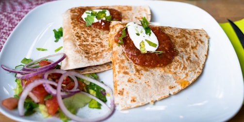 A serving of quesadillas is presented on a white plate, topped with salsa, sour cream, and garnished with cilantro. Beside the quesadillas, there is a small portion of salad with sliced red onions, tomatoes, and lettuce.