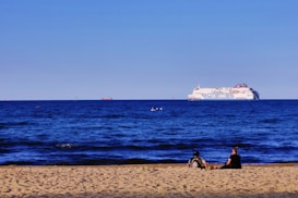 A vast blue sea stretches towards the horizon with a large ferry prominently visible in the distance. The words 'polferries' are displayed on its side. In the foreground, a sandy beach is occupied by two people sitting and enjoying the view. The sky is clear and light blue, adding to the serene atmosphere.