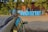 Large blue letters spelling out 'WARSZTAT' are positioned in front of a red brick building. The letters are on a sidewalk with trees and a chain-link fence in the background. A parked car is visible in the lower left corner of the image, and two people are standing on the right side.