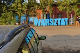 Large blue letters spelling out 'WARSZTAT' are positioned in front of a red brick building. The letters are on a sidewalk with trees and a chain-link fence in the background. A parked car is visible in the lower left corner of the image, and two people are standing on the right side.