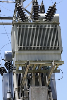 A close-up view of an electrical transformer mounted on a pole. The transformer has several insulators with spiral ridges arranged on top. Metal and ceramic components are part of the setup, and multiple wires are connected to the transformer. The clear blue sky is visible in the background.