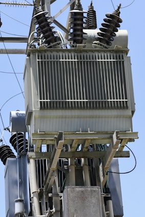 A close-up view of an electrical transformer mounted on a pole. The transformer has several insulators with spiral ridges arranged on top. Metal and ceramic components are part of the setup, and multiple wires are connected to the transformer. The clear blue sky is visible in the background.