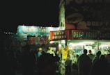 A nighttime carnival scene with a brightly lit food stall offering cold drinks and deep-fried candy. The stall is bustling with people gathered in front of it, and in the background, there is a brightly illuminated amusement park ride with flashing lights and colorful decorations.