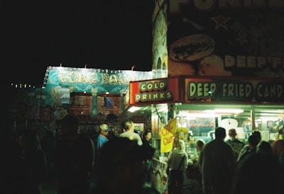 A nighttime carnival scene with a brightly lit food stall offering cold drinks and deep-fried candy. The stall is bustling with people gathered in front of it, and in the background, there is a brightly illuminated amusement park ride with flashing lights and colorful decorations.