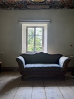 A vintage wooden sofa with blue patterned upholstery is positioned in front of a window with a scenic view of greenery outside. The room features an ornate, colorful ceiling pattern, adding a touch of elegance. The walls are painted in a muted tone, and the floor is made of wooden planks.