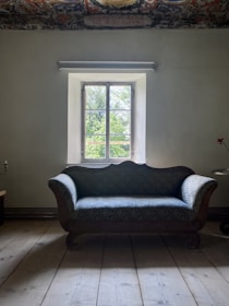 A vintage wooden sofa with blue patterned upholstery is positioned in front of a window with a scenic view of greenery outside. The room features an ornate, colorful ceiling pattern, adding a touch of elegance. The walls are painted in a muted tone, and the floor is made of wooden planks.
