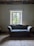 A vintage wooden sofa with blue patterned upholstery is positioned in front of a window with a scenic view of greenery outside. The room features an ornate, colorful ceiling pattern, adding a touch of elegance. The walls are painted in a muted tone, and the floor is made of wooden planks.