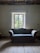 A vintage wooden sofa with blue patterned upholstery is positioned in front of a window with a scenic view of greenery outside. The room features an ornate, colorful ceiling pattern, adding a touch of elegance. The walls are painted in a muted tone, and the floor is made of wooden planks.