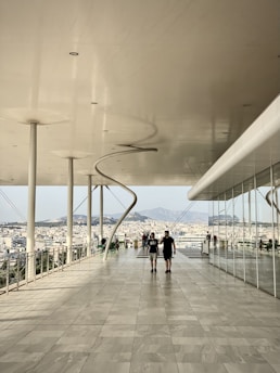 A modern, spacious terrace with a large overhang and tall supporting pillars. The floor is made of light-colored tiles. Two people are walking towards the camera in the middle of the terrace. In the background, there is a view of a city and distant mountains under a clear sky. Part of the building has a glass facade reflecting the outdoor scene.
