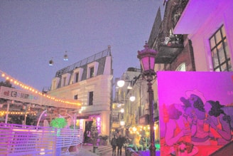 A lively street scene during the evening with a mix of architectural elements, including an old-style building with balconies and a modern structure adorned with string lights. There is vibrant street art featuring colorful illustrations of people with sunglasses and hats. The setting includes outdoor dining areas and decorative lighting creating a festive atmosphere.