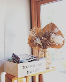 A wooden box labeled 'Drop Books Here Thank You' is placed on a wooden table next to a window. Inside the box, there are several children's books with colorful covers. Beside the box, there is a decorative vase containing dried leaves and pampas grass, adding a rustic touch to the arrangement.