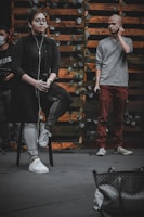 A person sits on a stool holding a microphone, wearing glasses, earbuds, a dark shirt, and jeans. Another person stands nearby, holding a microphone and adjusting an earbud, dressed in a gray sweater and red pants. The backdrop features wooden slats adorned with leafy vines, creating a rustic setting. Partially visible equipment and a bag are in the foreground.