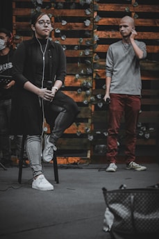 A person sits on a stool holding a microphone, wearing glasses, earbuds, a dark shirt, and jeans. Another person stands nearby, holding a microphone and adjusting an earbud, dressed in a gray sweater and red pants. The backdrop features wooden slats adorned with leafy vines, creating a rustic setting. Partially visible equipment and a bag are in the foreground.
