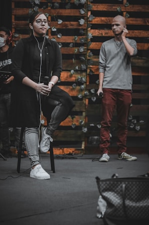 A person sits on a stool holding a microphone, wearing glasses, earbuds, a dark shirt, and jeans. Another person stands nearby, holding a microphone and adjusting an earbud, dressed in a gray sweater and red pants. The backdrop features wooden slats adorned with leafy vines, creating a rustic setting. Partially visible equipment and a bag are in the foreground.