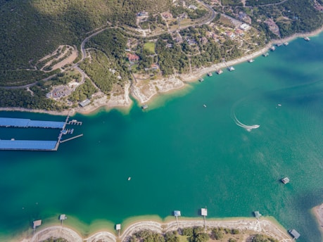 An aerial view of a large body of water surrounded by lush greenery and residential areas. Several docks extend into the water, and a boat is creating a curved wake on the surface. The shoreline features a mix of sandy and rocky areas.