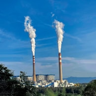 Two tall industrial smokestacks emitting white smoke stand against a clear blue sky. Below them is a large factory complex with visible structures and surrounding greenery.