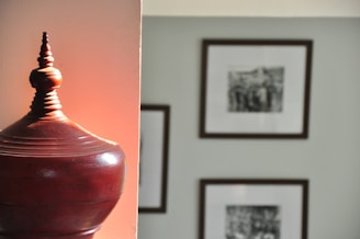 A wooden decorative object with a pointed top sits in the foreground with a warm, reddish hue cast on it. In the background, several black and white framed photographs are hung on a gray wall.