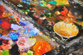 A vibrant array of mixed paints spread out on an artist's palette, showcasing a variety of colors including blue, red, orange, yellow, and green. A paint-smeared bowl is positioned to the side.