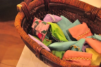 A woven wicker basket contains several small, colorful fabric pouches. The pouches are of different colors including pink, green, blue, and purple, and are embellished with intricate golden embroidery and patterns. One notable pouch has a unique design featuring cartoon-like characters.