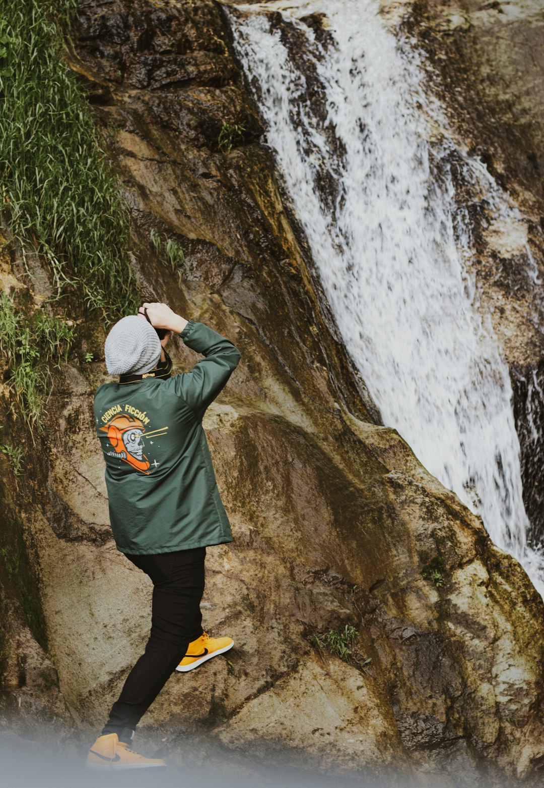 person in green jacket and black pants standing on brown rock near waterfalls during daytime