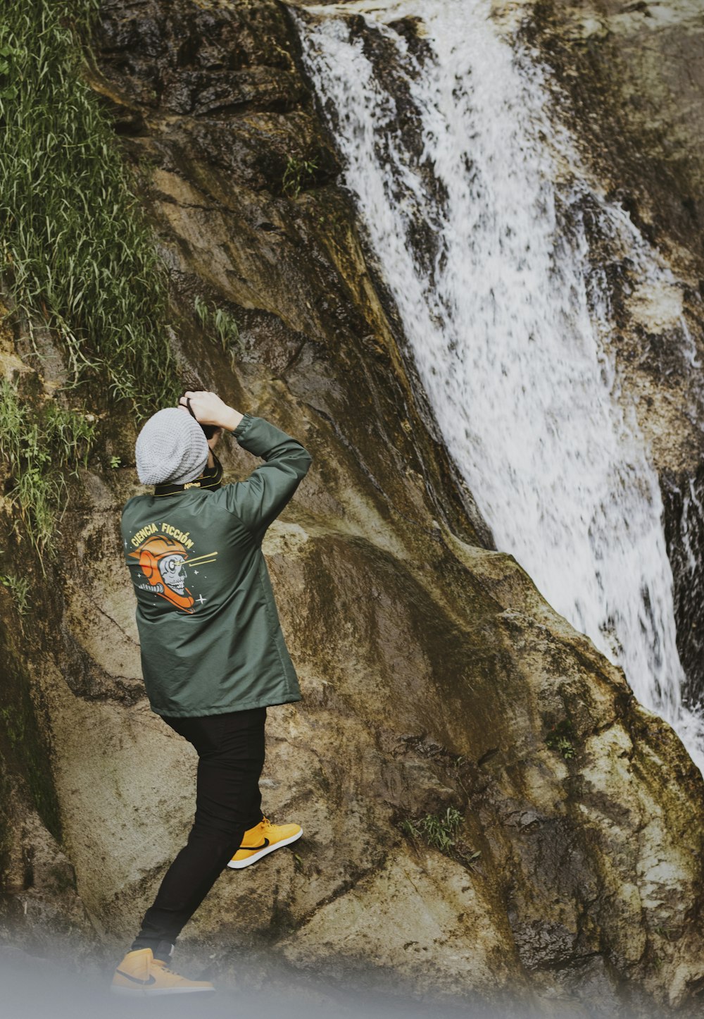 person in green jacket and black pants standing on brown rock near waterfalls during daytime