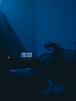 A dimly lit room with a blue color tone features a digital clock displaying 12:20. A bicycle is leaning against the wall, partially visible. The overall lighting creates a calm, subdued atmosphere.